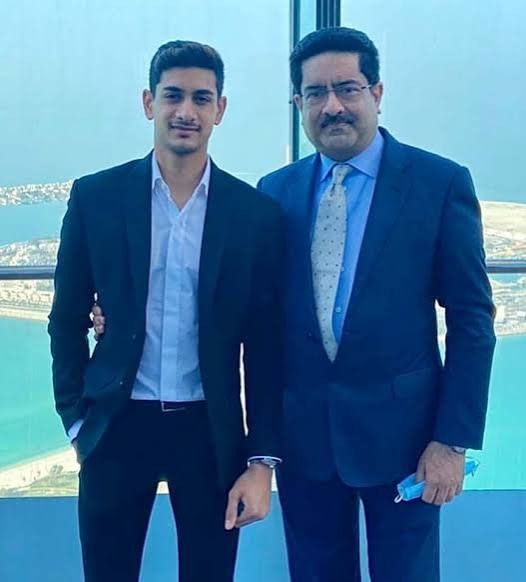 Aryaman Birla and his father Aditya Birla posing together with city skyline and water view behind them Two men in suits standing by window with modern cityscape and sea view Formal portrait of two men in business attire with urban waterfront background