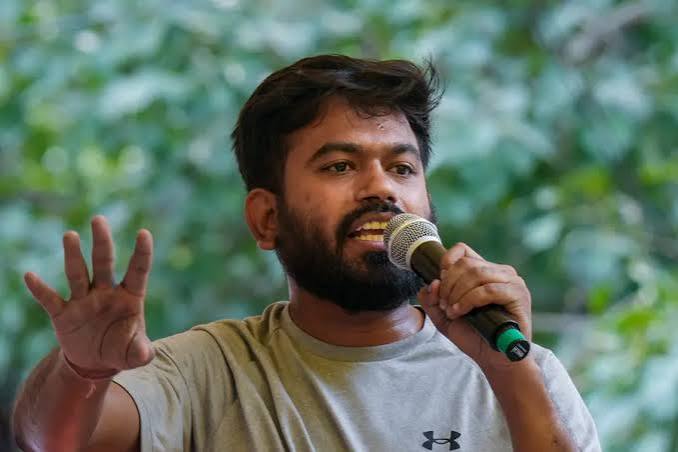 A portrait of Abhijeet Dipke wearing a grey Under Armour t-shirt, holding a microphone in his left hand and gesturing with an open palm in his right hand against a blurred green background.